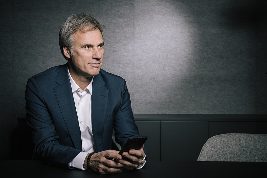 Bitkom-Präsident Achim Berg sitzt auf einer Bank und hält ein Smartphone in der Hand